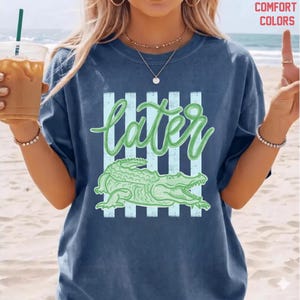 May include: Navy blue t-shirt with a green alligator graphic and the word "later" in a script font. The design includes vertical light blue stripes. The shirt is a comfort color.