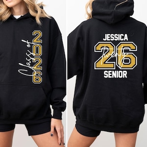 May include: Black hoodie with gold and white lettering. The front reads "Class of 2026" vertically. The back displays "JESSICA 26 SENIOR" in gold and white. Features a hood and a front pocket. A stylish and comfortable garment.
