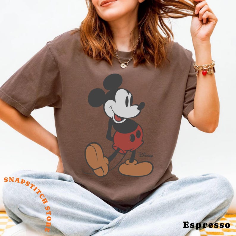 May include: Espresso-colored t-shirt featuring a graphic of Mickey Mouse in his classic pose. The cartoon character is depicted in black, white, and red. The shirt has the text "Disney" and "Snapstitch Store".