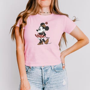 Może przedstawiać: Jasnor&oacute;żowa koszulka z grafiką Myszki Minnie. Postać z kresk&oacute;wki jest przedstawiona w czerwonej sp&oacute;dnicy w kropki, brązowych butach i pasującym kapeluszu. Logo Disneya jest wydrukowane poniżej.