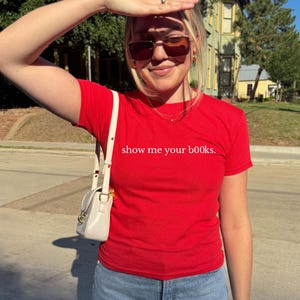 May include: A bright red t-shirt with the phrase "show me your books." printed in white. The shirt is paired with a small, off-white purse with a gold clasp. The person is wearing sunglasses.