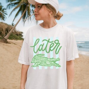 May include: White t-shirt with a green alligator graphic and the word "later" in a cursive font. The design includes light blue vertical stripes. The shirt is worn on a beach.