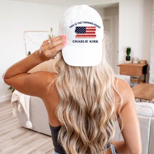 May include: White baseball cap with the text "THIS IS THE TURNING POINT" above a distressed American flag graphic and the name "CHARLIE KIRK" below. The cap is a classic six-panel design.