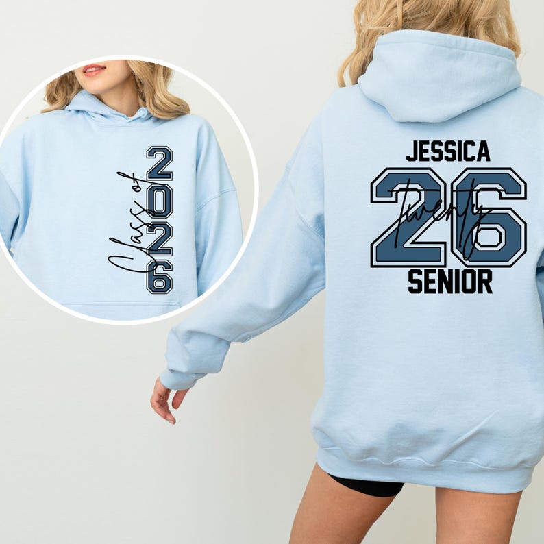 Pu&ograve; includere: Felpa con cappuccio azzurra con la scritta "Class of 2026" sul davanti e "Jessica 26 Senior" sul retro. La felpa ha un cappuccio con coulisse e maniche lunghe. Il testo &egrave; di colore blu scuro.
