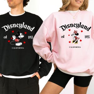 Pode incluir: Camisolas pretas e rosa com o log&oacute;tipo da Disneyland, gr&aacute;ficos do Mickey Mouse e da Minnie Mouse, e o texto "est 1955 CALIFORNIA". As camisolas t&ecirc;m mangas compridas e gola redonda.