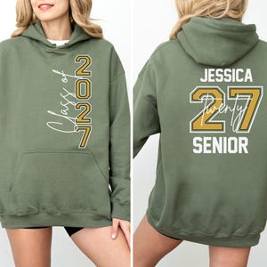 May include: Olive green hoodie with the text "Class of 2027" on the front and "Jessica Twenty 27 Senior" on the back. The hoodie has a hood and a front pocket. The text is in white and gold.