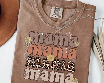 May include: A brown Comfort Colors t-shirt with the word "Mama" in a stacked design. The word "Mama" is in various colors and fonts, including a leopard print. The shirt also features floral and butterfly graphics. A silver necklace with a pendant is on the shirt.