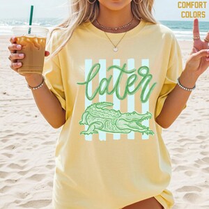 May include: Yellow t-shirt with a green alligator graphic and the word "later" in a script font. The shirt has a relaxed fit and is made of a soft material. The design includes vertical light blue stripes.