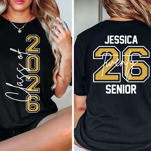 Puede incluir: Camiseta negra con letras doradas y blancas. La parte delantera muestra "Class of 2026" y la trasera dice "Jessica 26 Senior". Los n&uacute;meros est&aacute;n delineados en dorado con un borde blanco, y el nombre est&aacute; en escritura blanca.