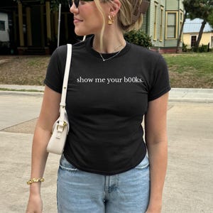 May include: Black crew neck t-shirt with the text "show me your books" in white. The shirt is paired with light blue jeans and a white purse. The person is wearing gold jewelry and sunglasses.