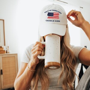 May include: White baseball cap with the text "THIS IS THE TURNING POINT" and "CHARLIE KIRK" below an American flag design. A large white mug with a brown base is held in front of the face.