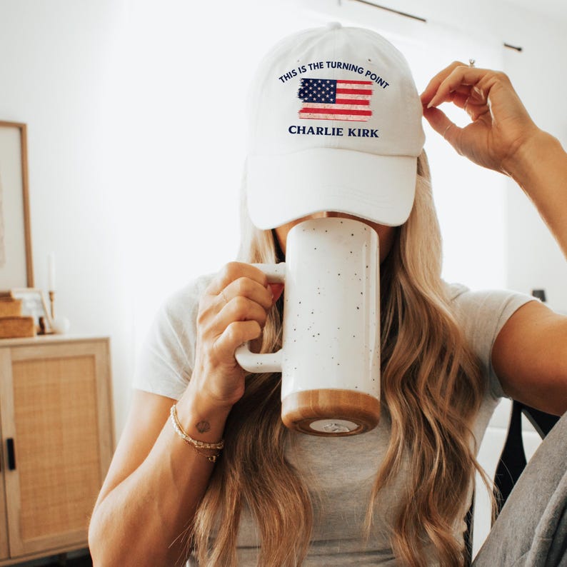 May include: White baseball cap with the text "THIS IS THE TURNING POINT" and "CHARLIE KIRK" below an American flag design. A large white mug with a brown base is held in front of the face.