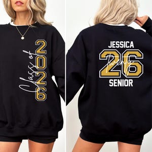 Pu&ograve; includere: Felpa nera con finiture bianche con "Class of 2026" e il numero "26" in oro e bianco. Il retro mostra "Jessica 26 Senior" in oro e bianco.