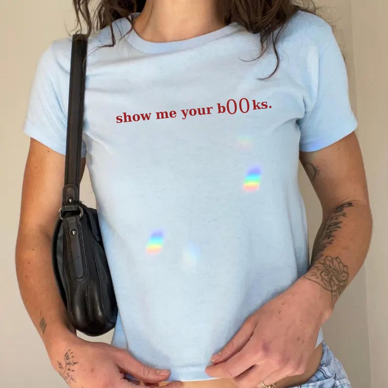 May include: Light blue t-shirt with the phrase "show me your books" in red lettering. The shirt is short-sleeved and appears to be made of a soft material. A black purse hangs from the shoulder.