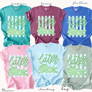 May include: A collection of six short-sleeved t-shirts in various colors, including teal, berry, and blue. Each shirt features the word "later" in a script font above a green alligator graphic, set against a striped background.