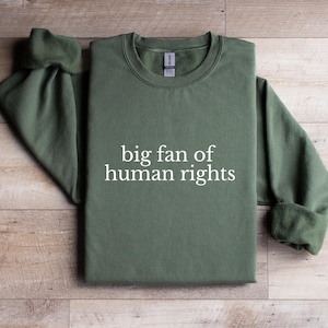 May include: An olive green sweatshirt with the phrase "big fan of human rights" printed in white. The sweatshirt has a crew neck and long sleeves with cuffed wrists. The garment is displayed on a light brown wooden surface.