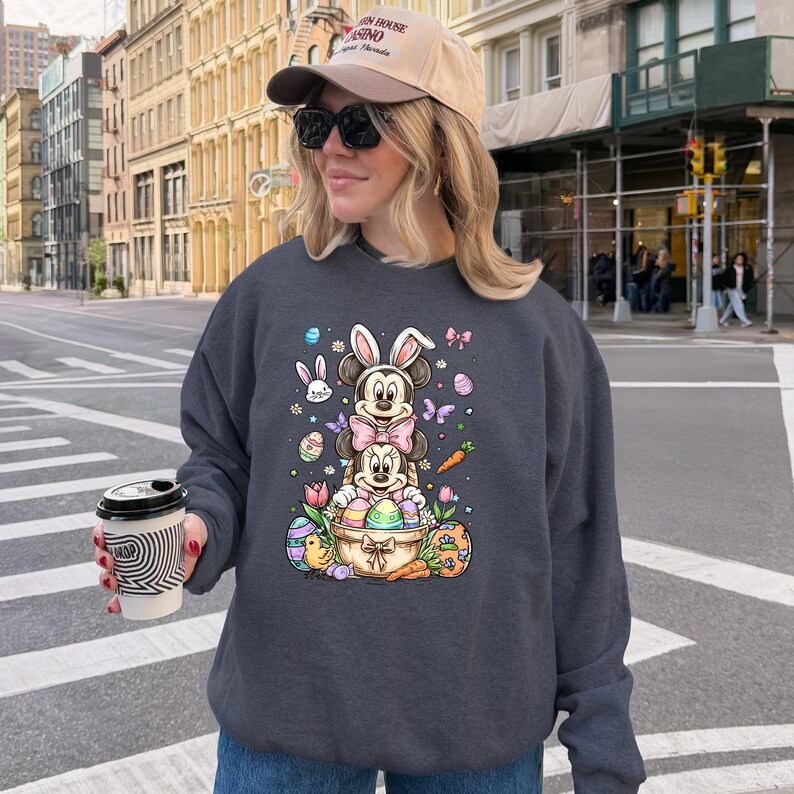 K&ouml;nnte beinhalten: Dunkelgraues Sweatshirt mit einem farbenfrohen Oster-Motiv. Das Motiv zeigt Mickey und Minnie Mouse-Figuren mit Hasenohren, Ostereiern, Karotten und Blumen. Das Sweatshirt hat einen Rundhalsausschnitt.