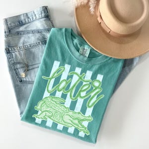 May include: A teal t-shirt with the word "later" in a cursive font and a green alligator graphic. The shirt is paired with light-wash denim jeans and a tan fedora hat. A gold necklace is visible.