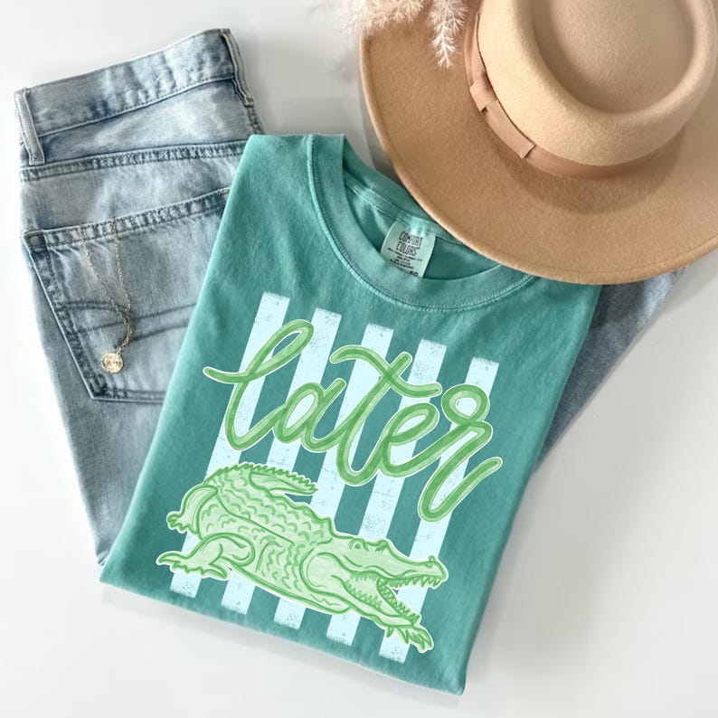 May include: A teal t-shirt with the word "later" in a cursive font and a green alligator graphic. The shirt is paired with light-wash denim jeans and a tan fedora hat. A gold necklace is visible.