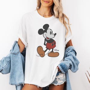 May include: White t-shirt featuring a classic Mickey Mouse graphic. The cartoon character is depicted in black, red, and brown, with the Disney logo printed on the lower right. The shirt has rolled-up sleeves.