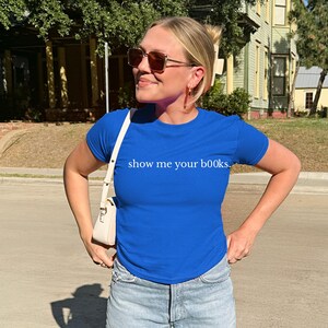 May include: A royal blue t-shirt with the text "show me your books." in white. The shirt is short-sleeved and form-fitting. The person is wearing sunglasses and a white purse.