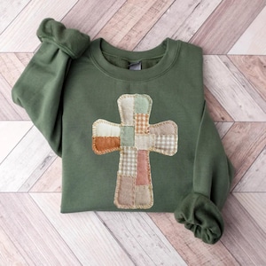 May include: Olive green sweatshirt featuring a patchwork cross design. The cross is made of various fabric squares in neutral tones, including beige, brown, and green. The sleeves have a gathered cuff detail. The sweatshirt is laid flat on a light wood-grain surface.