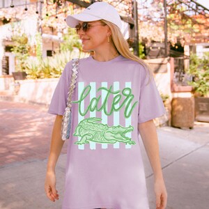 May include: A lilac-colored t-shirt with the word "later" in green script above a green alligator graphic. The word and alligator are set against a backdrop of vertical light blue stripes.
