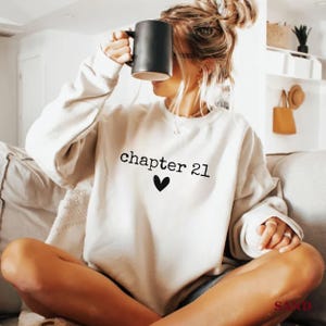 Peut inclure: Un sweat-shirt crème avec le texte "chapter 21" au-dessus d'un cœur noir. Le sweat-shirt est de style col rond. Une tasse noire est tenue près du visage.