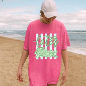 May include: A hot pink t-shirt with a green alligator graphic and the word "later" in a cursive font. The design includes vertical light blue stripes. The shirt is worn on a beach.