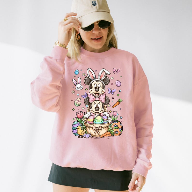 K&ouml;nnte beinhalten: Ein hellrosa Sweatshirt mit einem farbenfrohen Ostermotiv. Das Motiv zeigt die Charaktere Mickey und Minnie Mouse mit Hasenohren, umgeben von Ostereiern, Karotten und Schmetterlingen. Das Design wirkt verspielt und festlich.