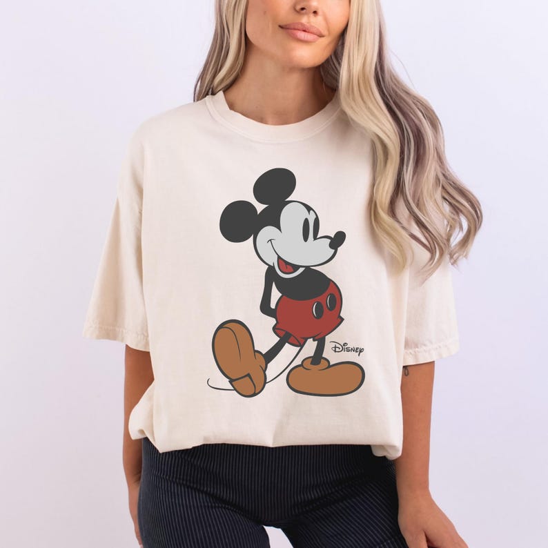 May include: Cream-colored t-shirt featuring a classic Mickey Mouse graphic. The cartoon character is depicted in his iconic pose, wearing red shorts, brown shoes, and black gloves. The Disney logo is printed on the bottom right of the graphic.
