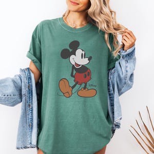 May include: Green t-shirt featuring a classic Mickey Mouse graphic. The cartoon character is depicted in black, white, red, and brown, with the Disney logo below. The shirt has short sleeves and a relaxed fit.