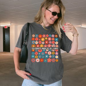 May include: A dark grey t-shirt with a vibrant floral pattern and the words "CELEBRATE MINDS OF ALL KINDS". The design features flowers in red, orange, blue, and yellow. The t-shirt has a loose fit and short sleeves.
