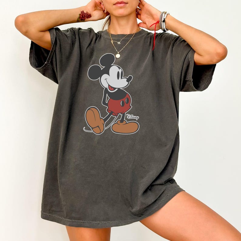 May include: Dark gray t-shirt featuring a classic Mickey Mouse graphic. The cartoon character is depicted in black, red, white, and brown, with the Disney logo. The shirt has short sleeves and a relaxed fit.