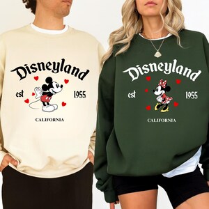 Pode incluir: Duas camisolas, uma creme e outra verde escuro, com o log&oacute;tipo da Disneyland e Mickey e Minnie Mouse. O texto inclui "est 1955" e "CALIFORNIA". Mickey usa cal&ccedil;&otilde;es vermelhos, Minnie um la&ccedil;o de bolinhas vermelhas.