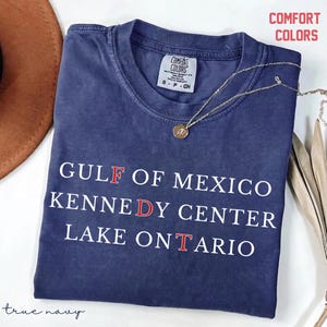 May include: A navy blue short-sleeve t-shirt with white text that reads GULF OF MEXICO, KENNEDY CENTER, and LAKE ONTARIO, with the letters F, D, and T highlighted in red. The shirt features a Comfort Colors brand label at the inner neckline.
