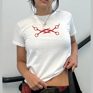 May include: White t-shirt featuring a red graphic of crossed scissors. The short-sleeved shirt is made of a soft material. The design is centered on the chest. The shirt is a casual style.