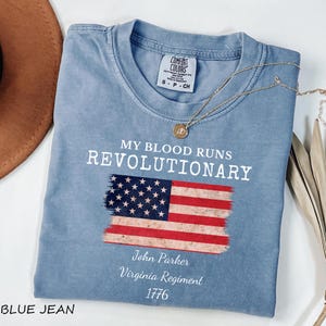 May include: A blue jean colored t-shirt with the text "MY BLOOD RUNS REVOLUTIONARY" and an American flag graphic. The shirt also includes the text "John Parker, Virginia Regiment, 1776". A gold necklace is draped across the shirt.