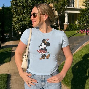 Może przedstawiać: Jasnoniebieska koszulka z motywem Myszki Minnie. Postać z kresk&oacute;wki ma na sobie czerwoną sp&oacute;dnicę w kropki i brązowe buty. Koszulka ma kr&oacute;tkie rękawy i okrągły dekolt. Logo Disneya jest wydrukowane poniżej.