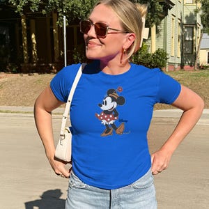 Może przedstawiać: Kr&oacute;lewski niebieski t-shirt z grafiką Myszki Minnie. Postać z kresk&oacute;wki ma na sobie sp&oacute;dnicę w kropki, kapelusz i buty. Koszulka ma okrągły dekolt i kr&oacute;tkie rękawy. Logo Disneya jest wydrukowane w prawym dolnym rogu.