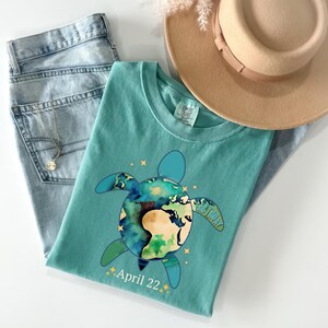 May include: A turquoise t-shirt featuring a turtle design with the Earth as its shell. The shirt includes the text "April 22." and is paired with light-wash jeans and a tan hat. A gold necklace is visible.