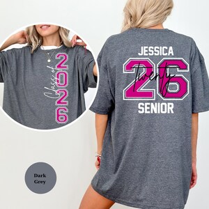 Puede incluir: Camiseta gris oscuro con el texto "Class of 2026" en un dise&ntilde;o vertical en la parte delantera. La parte trasera de la camiseta presenta el nombre "Jessica" encima del n&uacute;mero "26" en un dise&ntilde;o rosa y blanco, con la palabra "SENIOR" debajo.