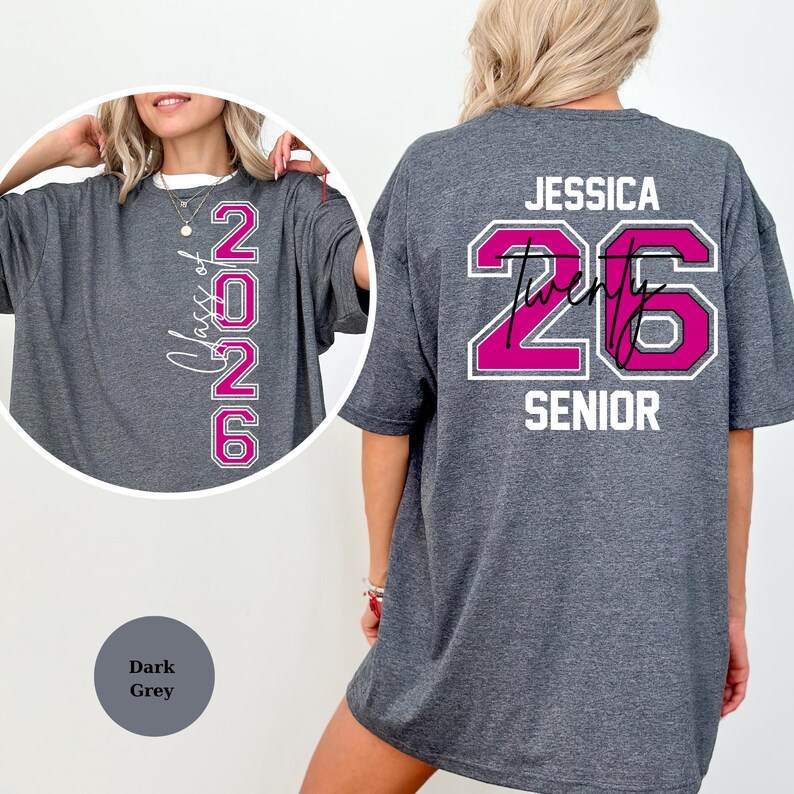 Puede incluir: Camiseta gris oscuro con el texto "Class of 2026" en un dise&ntilde;o vertical en la parte delantera. La parte trasera de la camiseta presenta el nombre "Jessica" encima del n&uacute;mero "26" en un dise&ntilde;o rosa y blanco, con la palabra "SENIOR" debajo.