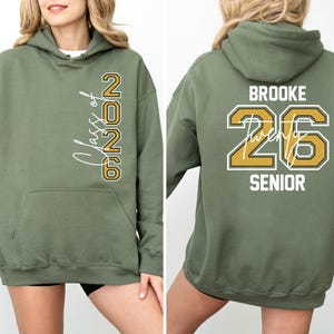Pu&ograve; includere: Felpa con cappuccio verde oliva con le scritte "Class of 2026" e "Brooke 26 Senior" stampate in bianco e oro. La felpa ha una tasca frontale e un cappuccio. Un capo casual per la laurea.