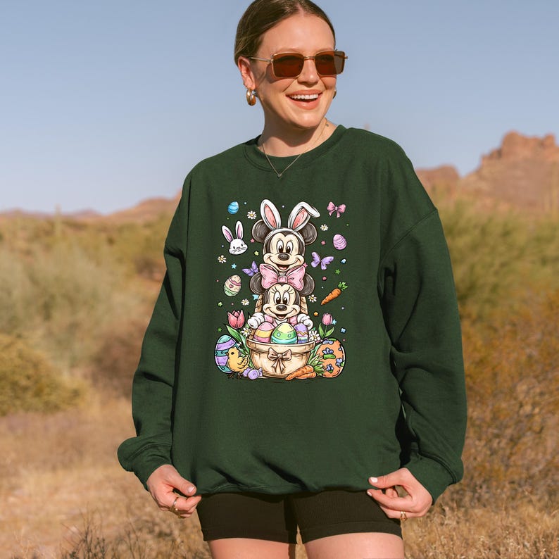 K&ouml;nnte beinhalten: Dunkelgr&uuml;nes Sweatshirt mit einem Ostermotiv. Das Design zeigt Mickey- und Minnie-Mouse-Figuren mit Hasenohren, umgeben von Ostereiern, Schmetterlingen und Karotten. Das Sweatshirt wird von einer Person getragen.