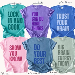May include: Six brightly coloured t-shirts with motivational slogans. The shirts are seafoam, violet, flo blue, blossom, blue jean, and bay. Each shirt features a different phrase in a contrasting colour, such as "Lock in and Cook" and "Trust Your Brain."