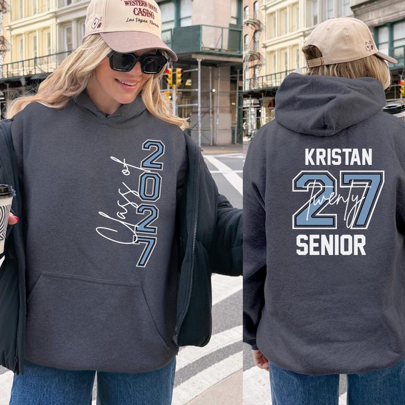 May include: Dark gray hoodie with a front pocket and a hood. The front features the text "Class of 2027" in a script font with a light blue outline. The back displays "Kristan 27 Senior" in a similar style.