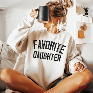 May include: Cream-colored sweatshirt with the text "FAVORITE DAUGHTER" in bold black letters. A black mug is held up near the face. The sweatshirt is worn while sitting on a couch.