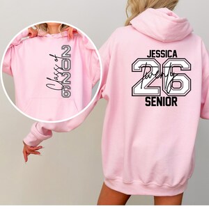 Pu&ograve; includere: Una felpa con cappuccio rosa chiaro con la scritta "Class of 2026" sul davanti e "Jessica Twenty 26 Senior" sul retro in bianco e nero. La felpa ha un cappuccio e una tasca frontale.