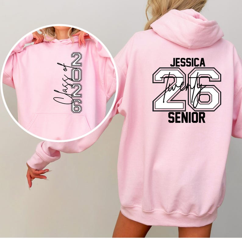 Pu&ograve; includere: Una felpa con cappuccio rosa chiaro con la scritta "Class of 2026" sul davanti e "Jessica Twenty 26 Senior" sul retro in bianco e nero. La felpa ha un cappuccio e una tasca frontale.